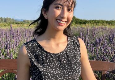 Photo of Tatiana sitting on a bench in front of a field of lavender. She is wearing a dark shirt with a flower print, and is smiling. Tatiana Benevides